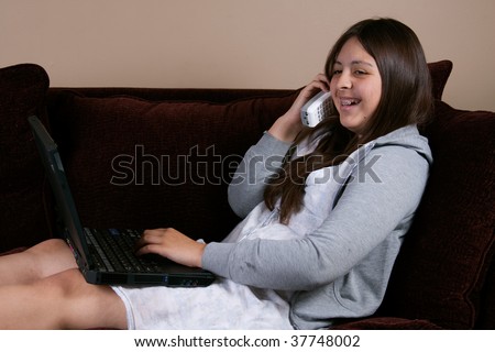 Pretty brunette hispanic teenage girl with braces wearing pajamas sitting on sofa or couch typing on laptop and talking on phone laughing