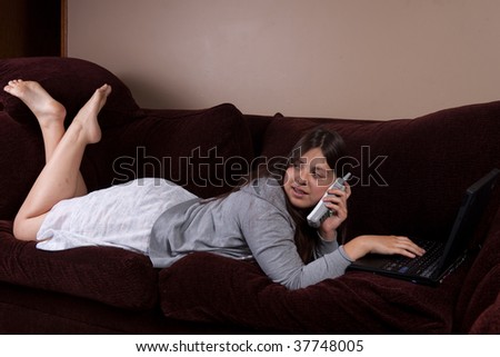 Pretty brunette hispanic teenage girl wearing pajamas laying on sofa or couch typing on laptop and talking on phone and looking at television