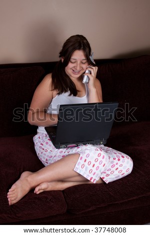 Pretty brunette hispanic teenage girl wearing pajamas sitting on sofa or couch typing on laptop and talking on phone