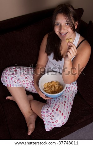 Pretty brunette teenage girl with braces sitting crossed legged on a sofa or couch eating a bowl of potato chips