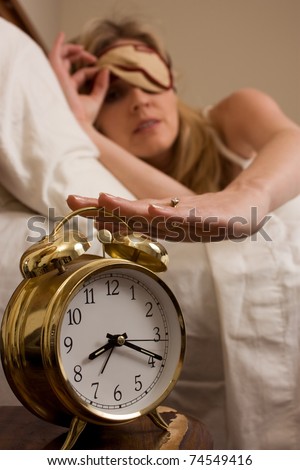 Blond woman sleeping in bed peeking out from cover over eyes looking at the clock and reaching to turn off a round gold alarm clock time is after eight o\'clock