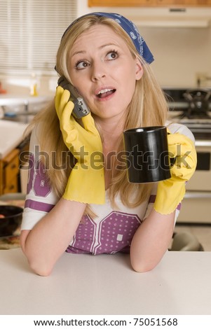 Blond caucasian woman wearing yellow cleaning gloves and hair scarf holding a cup of coffee talking on phone while leaning on a kitchen counter