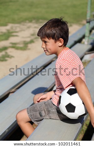 Young boy watches a sports game from the bleachers with a soccer ball under his arm