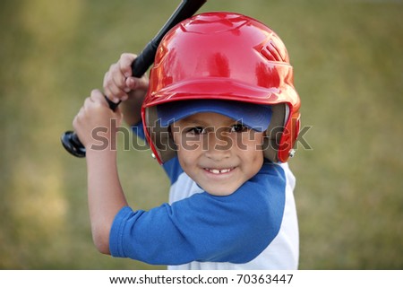Young hispanic or latino boy with red baseball helmet over a blue hat and blue tee shirt.
