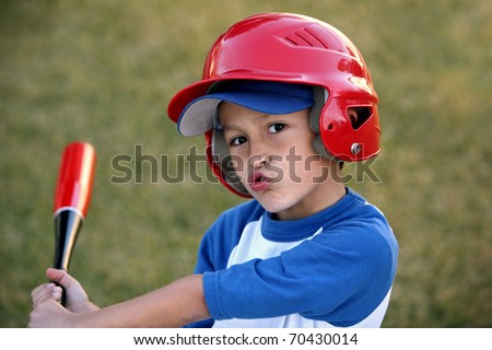 Young hispanic or latino boy with red baseball helmet over a blue hat and blue tee shirt.