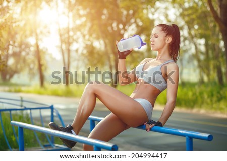 fitness woman relax after workout exercises on parallel bars outdoor with shaker bottle of water. brunette fit girl relax after street training