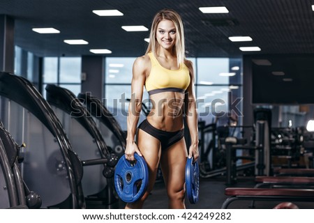 Sexy athletic sport smiling girl with perfect fitness body doing workout hard training with weights in the gym