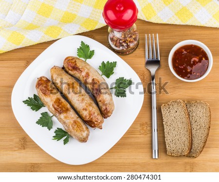 Fried pork sausages with parsley in glass plate, pepper, fork, bread and ketchup on wooden bamboo table, top view