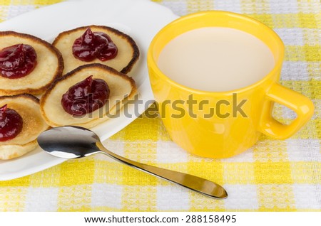 Pancakes with raspberry jam in glass dish and cup of milk on yellow tablecloth