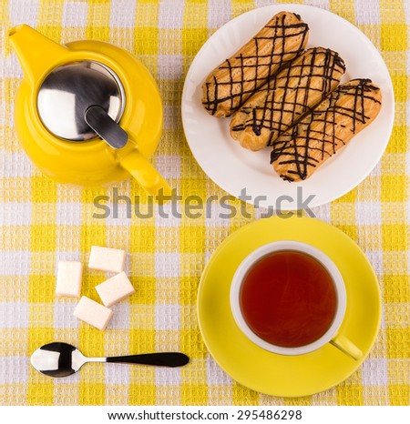 Hot tea in cup, sugar cubes and eclairs on yellow plaid tablecloth, top view