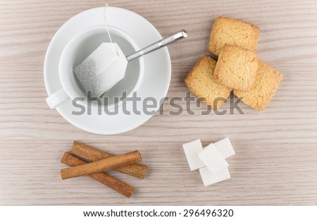 Cup with tea bag, cinnamon sticks, shortbread cookie and pieces of sugar on wooden table, top view
