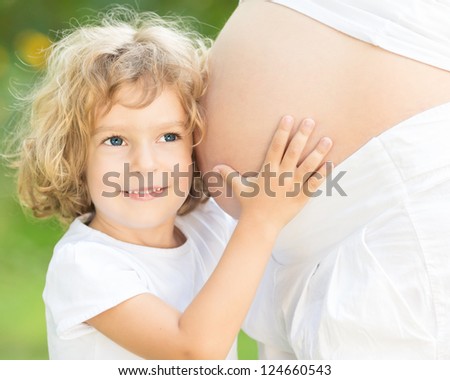 Happy child listen belly of pregnant woman against green spring background