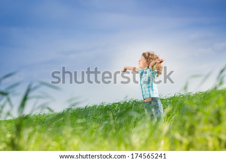 Happy kid with raised arms in green spring field against blue sky. Freedom and happiness concept