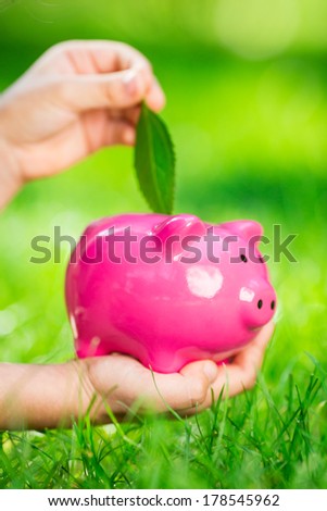 Piggybank and leaf in hands against green spring background. Shallow depth of field