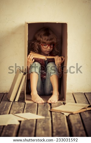 Child sitting in cardboard box. Kid reading book. Freedom and imagination concept. Unusual portrait