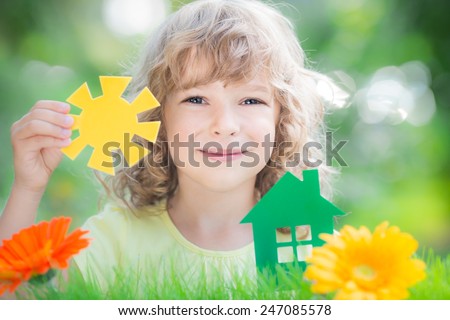Child holding green paper house and sun in hands against spring background. Real estate concept