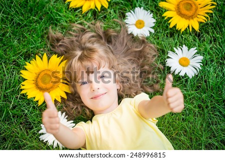 Happy child lying on grass. Funny kid playing in park. Beautiful spring flowers. Unusual top view portrait. Success and winner. Thumbs up. Like