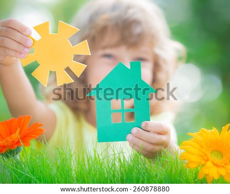 Happy child holding house and sun in hands against spring green background. Real estate business concept