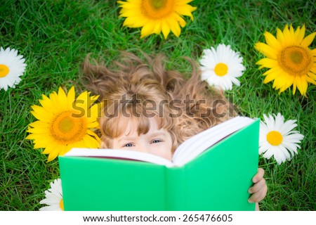 Happy child lying on green grass. Funny kid reading the book in spring park