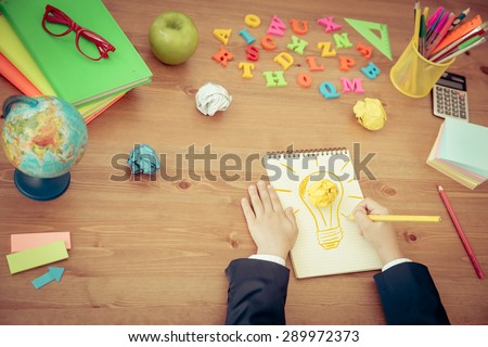 Child drawing lamp bulb in copybook. School items on wooden desk in class. New bright idea concept. Top view