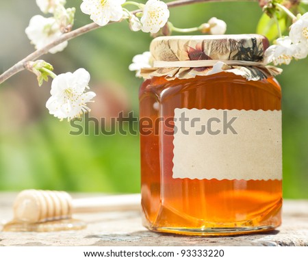 Honey jar with blank paper label, flower and wooden stick on table against green spring natural background