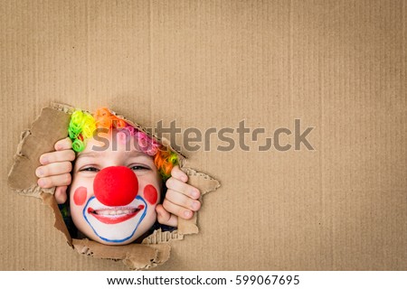 Funny kid clown looking through hole on cardboard. Child playing at home. 1 April Fool\'s day concept. Copy space.
