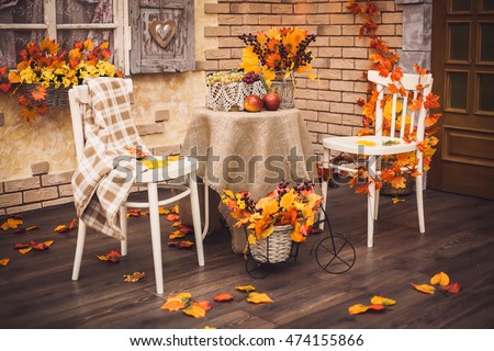 A cozy patio. Autumn leaves lying on the wooden floor, at the center are two white chairs and a table with burlap, fruits and a vase of berries and foliage. Background is brick wall and vintage window