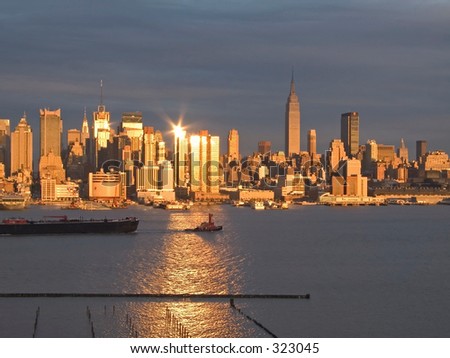 A tugboat pulls a barge along the Hudson River as the sunset reflects off the New York City skyline.