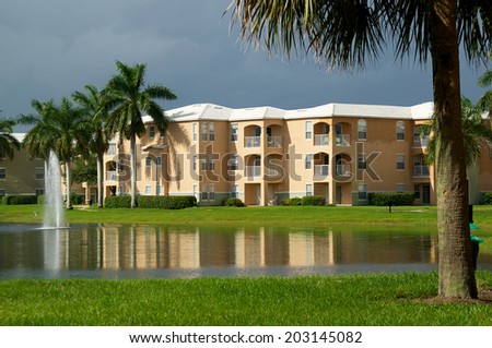 Looking across a pond towards a three story apartment complex in Naples, Florida, with a stormy sky and sunlight on buildings.