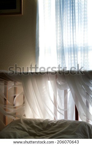 A simple bedroom under ambient light from the window coming in through blue sheer curtains to light up the sheer fabric wrapped around the headboard of the bed.