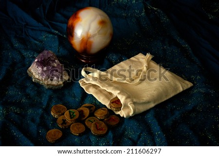 A plain muslin bag with wooden runes spilling out over a sparkling blue cloth with amethyst and crystal ball in the background.