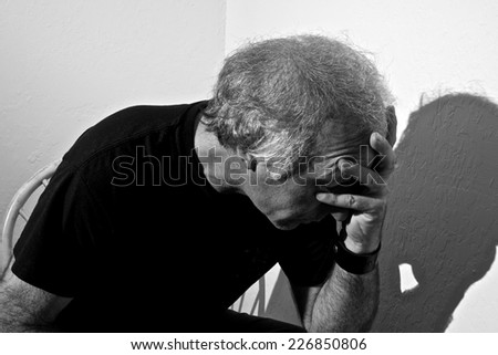 Portrait of a distressed older white man holding head sitting in corner of white room.