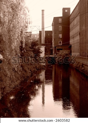 sepia image of canal behind old factory building in easthampton massachusetts, on the left is a bank of trees, on the right a bank of factory windows, in the center a smoke stack