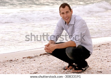 A casually well dressed smiling young adult crouches at the water's edge at the beach.