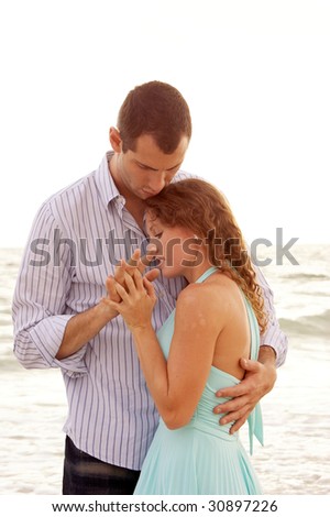 A woman being held close by her man, they are holding hands and they look like they are having a private with ocean waves in the background.