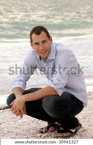 Handsome portrait of young man crouching on the beach , smiling and making eye contact with viewer