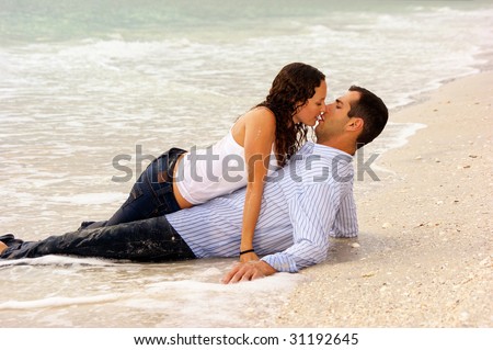 two lovers on the beach, laying in the water, the woman is dripping wet and laying on top of the man about to give him a kiss.