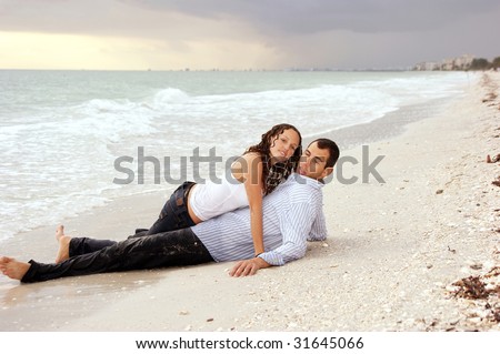 A beautiful you lady is laying on top of a man at the beach, their clothes are wet and she is looking at viewer, he has strange expression on his face at sunset.