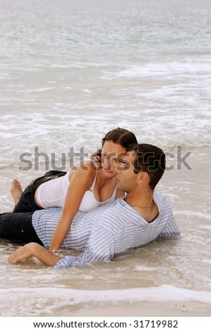 Beautiful young couple laying in the water at the beach, on top of each other, fully clothed. Heads touching looking to the left. With copy space on top.