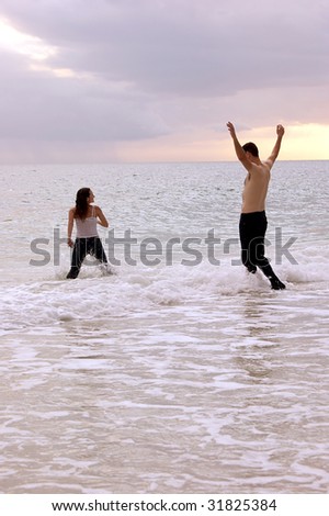 a woman appears to be running into the ocean followed by a tall man, shirtless with his arms up, they are playing in the water at sunset.