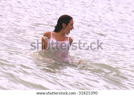 A fully clothed wet young woman in the middle of the sea has a surprised and happy look on her face with water splashing.