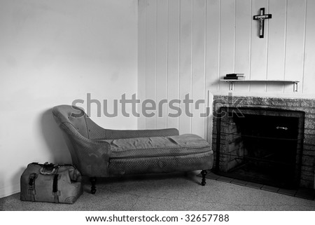 run down room with old sofa and travel bag. There is also a fireplace and a crucifix hanging above the shelf which has two books on it.