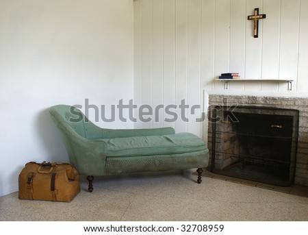 run down room with old sofa and travel bag. There is also a fireplace and a crucifix hanging above the shelf which has two books on it.
