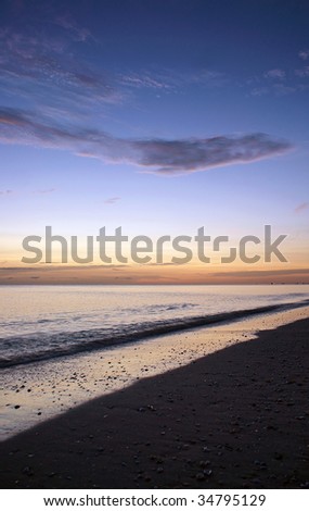 a rich blue sky fades to various colors on the horizon as the sun sets and the tide comes in on the beach