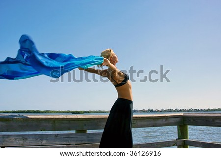 a beautiful fit woman is stretching in the sunshine, standing tall with her arms reaching out behind her with blue silk blowing in the breeze, plenty of room for copy or text