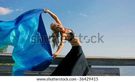 attractive fit blonde woman looking towards viewer smiling is leaning back holding her arms up and letting a beautiful silk cloth flow through the air behind her with room for copy or text in blue sky
