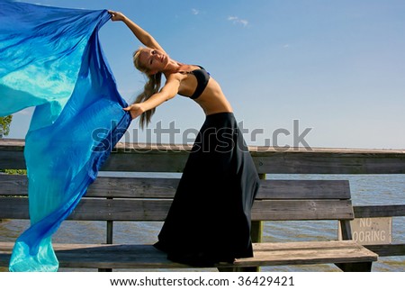 attractive fit blonde woman looking towards viewer is leaning back holding her arms up and letting a beautiful silk cloth flow through the air behind her against a blue sky with room for copy or text