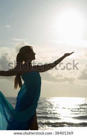 a blonde woman in profile standing at the water in a sheer dress is reaching up to catch the sun in her hand