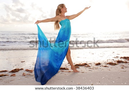 a female dancer is posing in profile with arms raised and looking at her open hand at the beach with waves crashing in the background