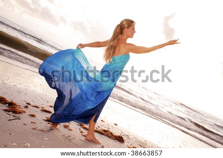 a female dancer is posing in profile with arms raised and looking at her open hand at the beach with waves crashing in the background, in a statuesque goddess like pose.